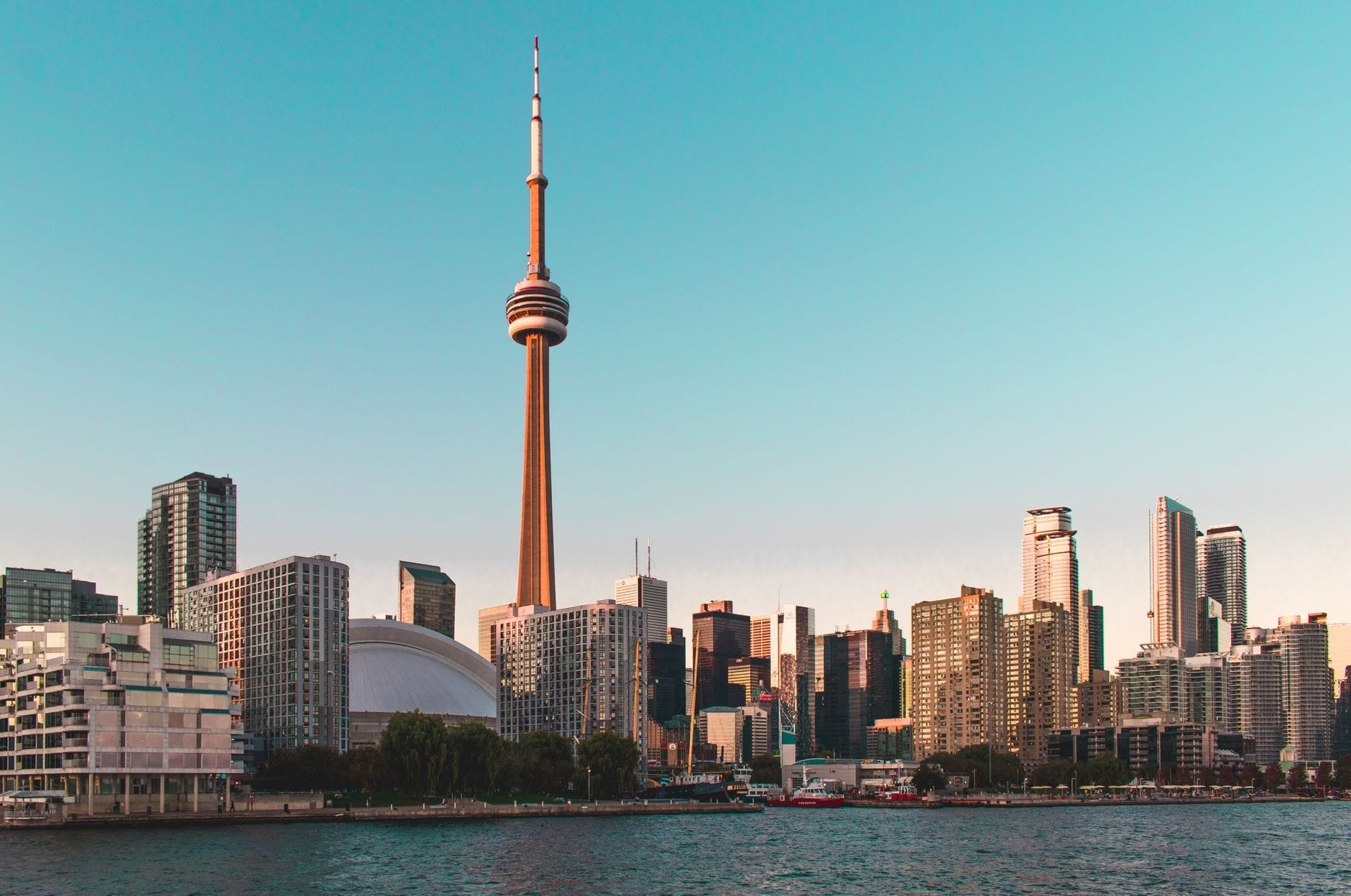 Skyline de Toronto ao pôr do sol com a CN Tower, o Rogers Centre e arranha-céus modernos, com iluminação quente e o Lago Ontário em primeiro plano.