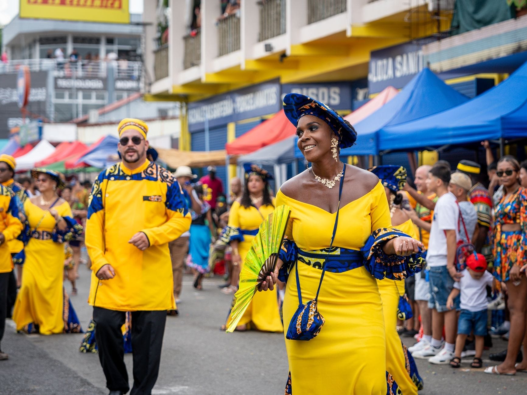 Participantes fantasiados com roupas amarelas vibrantes dançam e sorriem enquanto desfilam por uma rua da cidade, ladeada por tendas coloridas e espectadores durante um animado festival.
