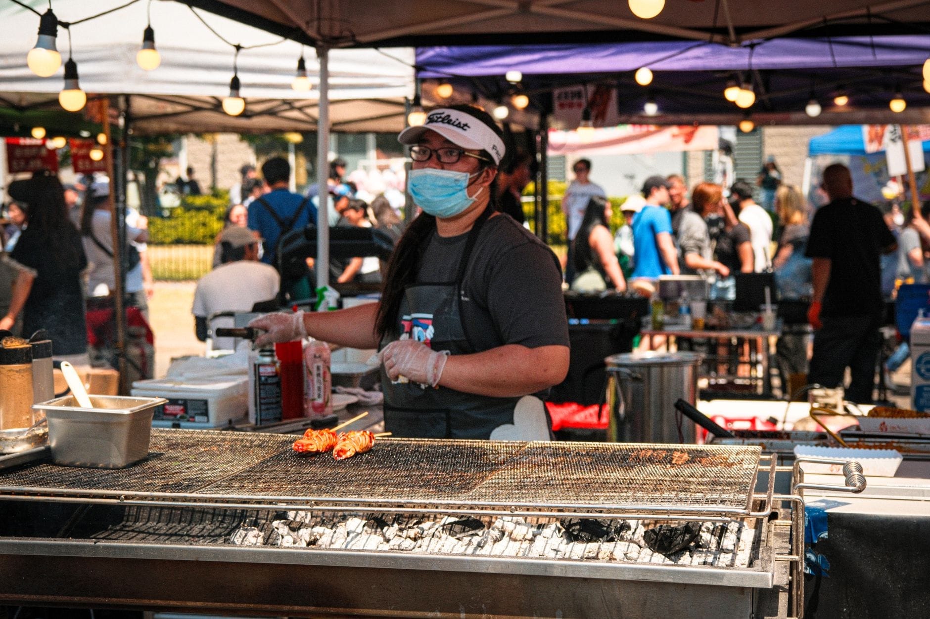 Vendedor de comida em festival ao ar livre grelhando espetinhos com uma multidão ao fundo em um animado evento de verão.