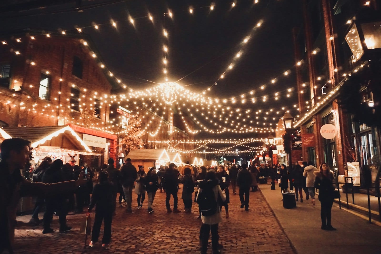 ผู้คนเดินตามถนนหินในย่าน Distillery District ของโตรอนโตยามค่ำ ท่ามกลางไฟประดับวันหยุดและซุ้มตลาดไม้