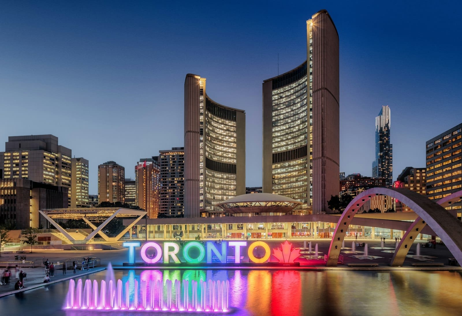 ศาลากลางโตรอนโตยามค่ำ พร้อมหอคู่งอแสง สัญลักษณ์ Toronto สีสันสด และน้ำพุที่ Nathan Phillips Square