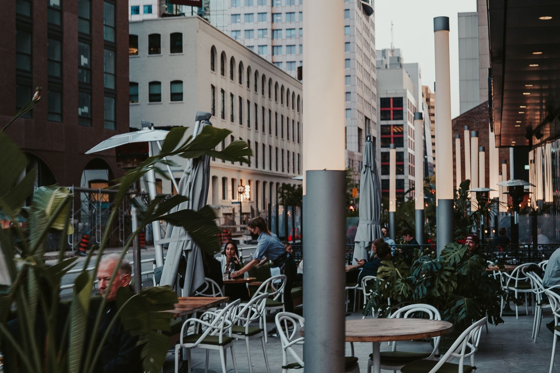 Pátio de rua no centro de Toronto com clientes em mesas rodeadas de edifícios modernos e luzes da cidade ao entardecer.