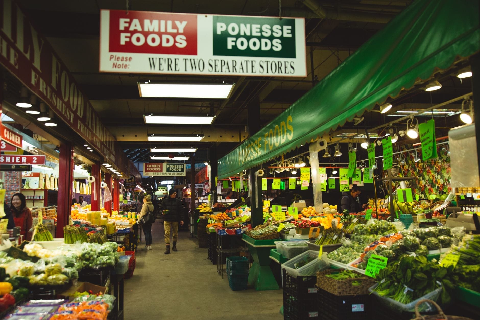 Mercado alimentar coberto com bancas de produtos frescos, legumes e frutas, compradores a explorar e cartazes suspensos numa rua movimentada.