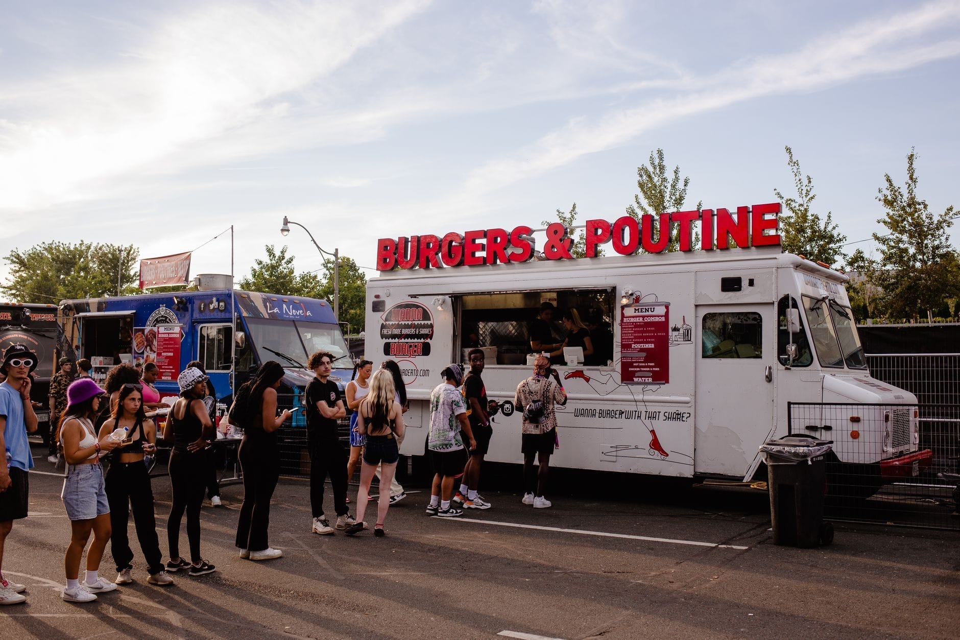 Pessoas aguardam na fila nos food trucks de Toronto, incluindo um caminhão branco com uma placa vermelha brilhante de 'Burgers & Poutine', sob um céu azul.