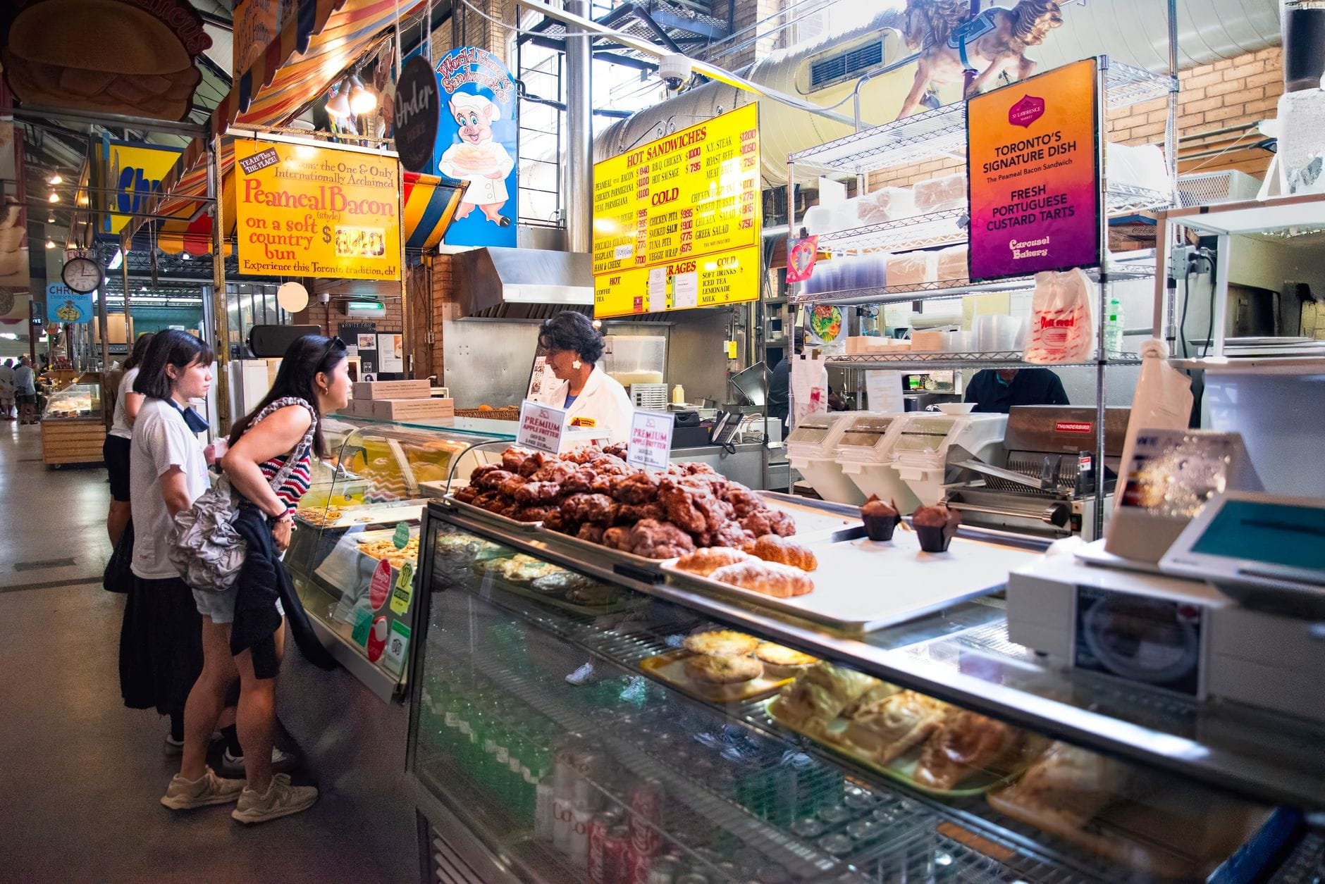Pessoas interagindo com um vendedor em um mercado de alimentos interno em Toronto, com pratos prontos, pães e doces, e placas coloridas acima do balcão.