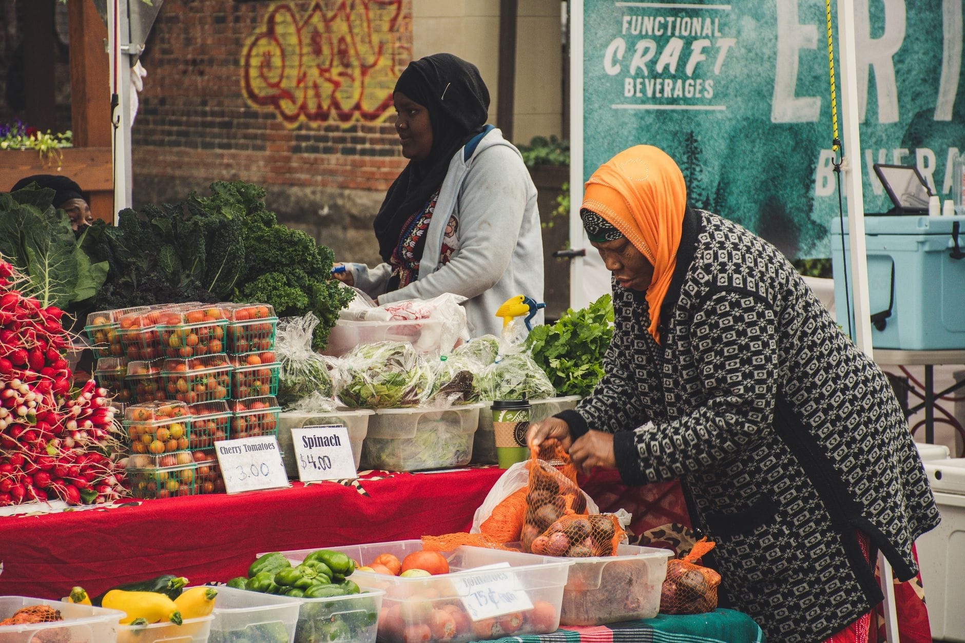 Duas mulheres vendendo produtos frescos, incluindo tomates, verduras e pimentões, em uma barraca de mercado comunitário com placas escritas à mão.