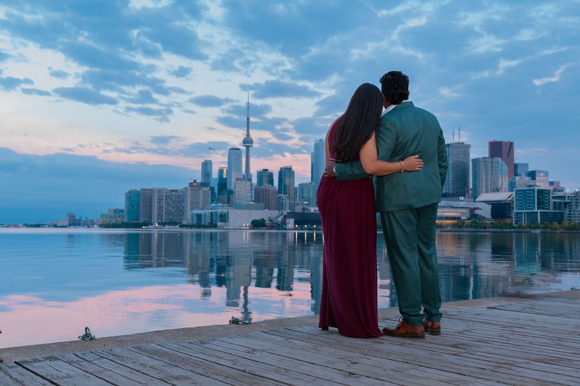 Casal se abraçando à beira da orla de Toronto ao entardecer, com o skyline e a CN Tower ao fundo refletidos nas águas calmas sob um céu azul dramático.