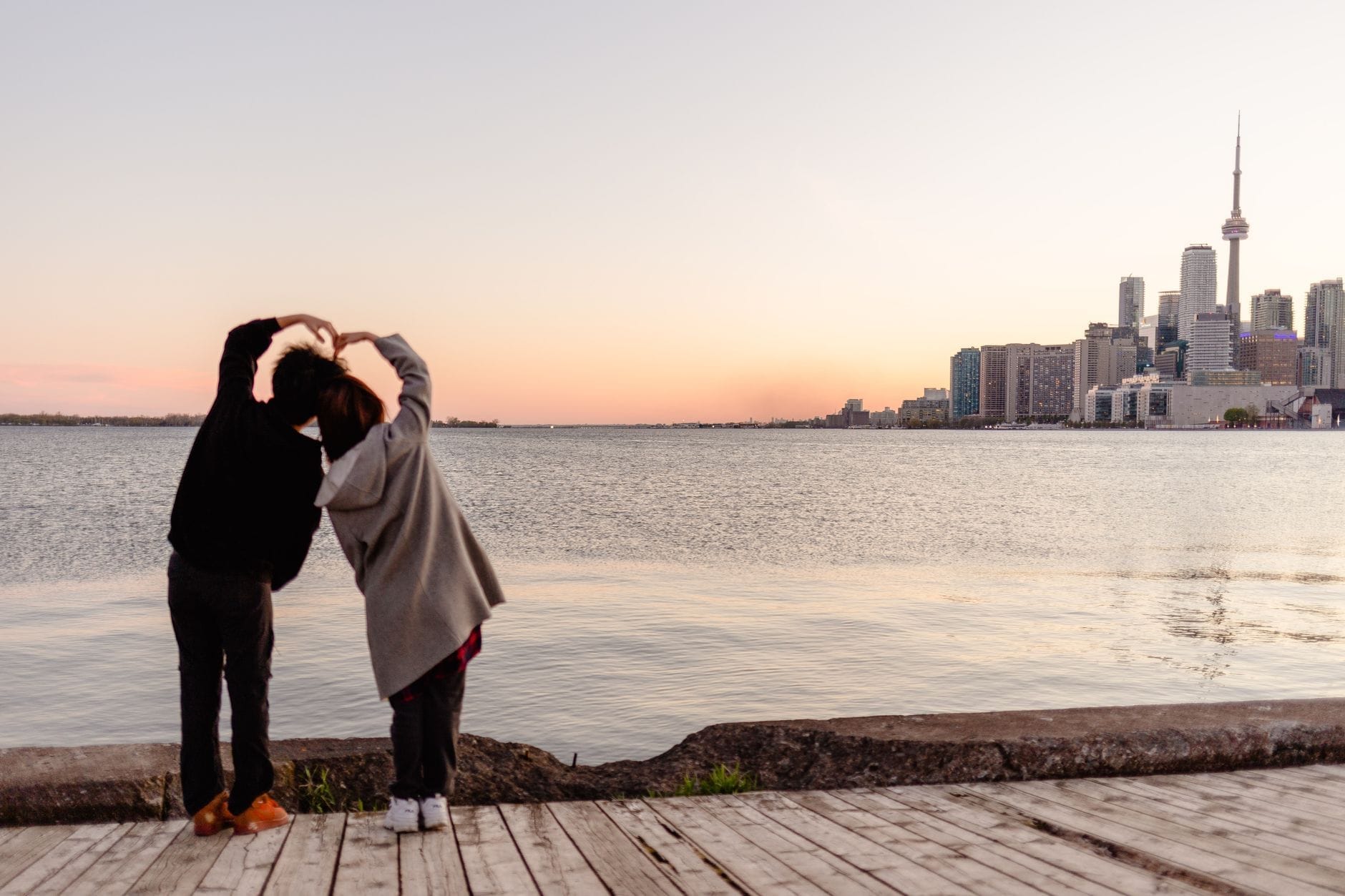 Casal fazendo formato de coração com os braços em frente ao lago Ontario com o skyline de Toronto ao pôr do sol