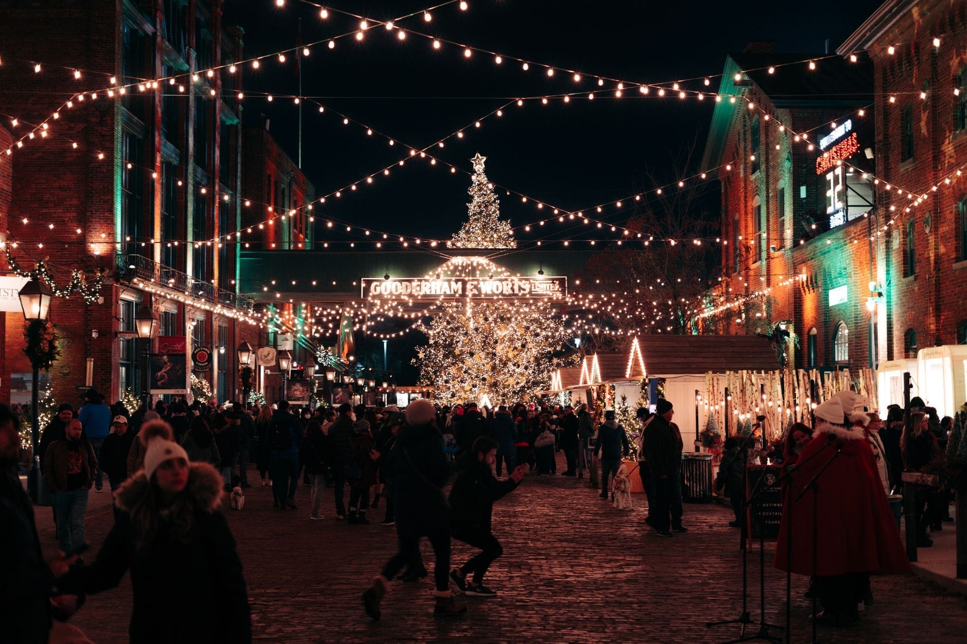 Multidões aproveitam uma noite de inverno festiva no Distillery District de Toronto, com luzes, barracas de mercado e uma grande árvore de Natal.