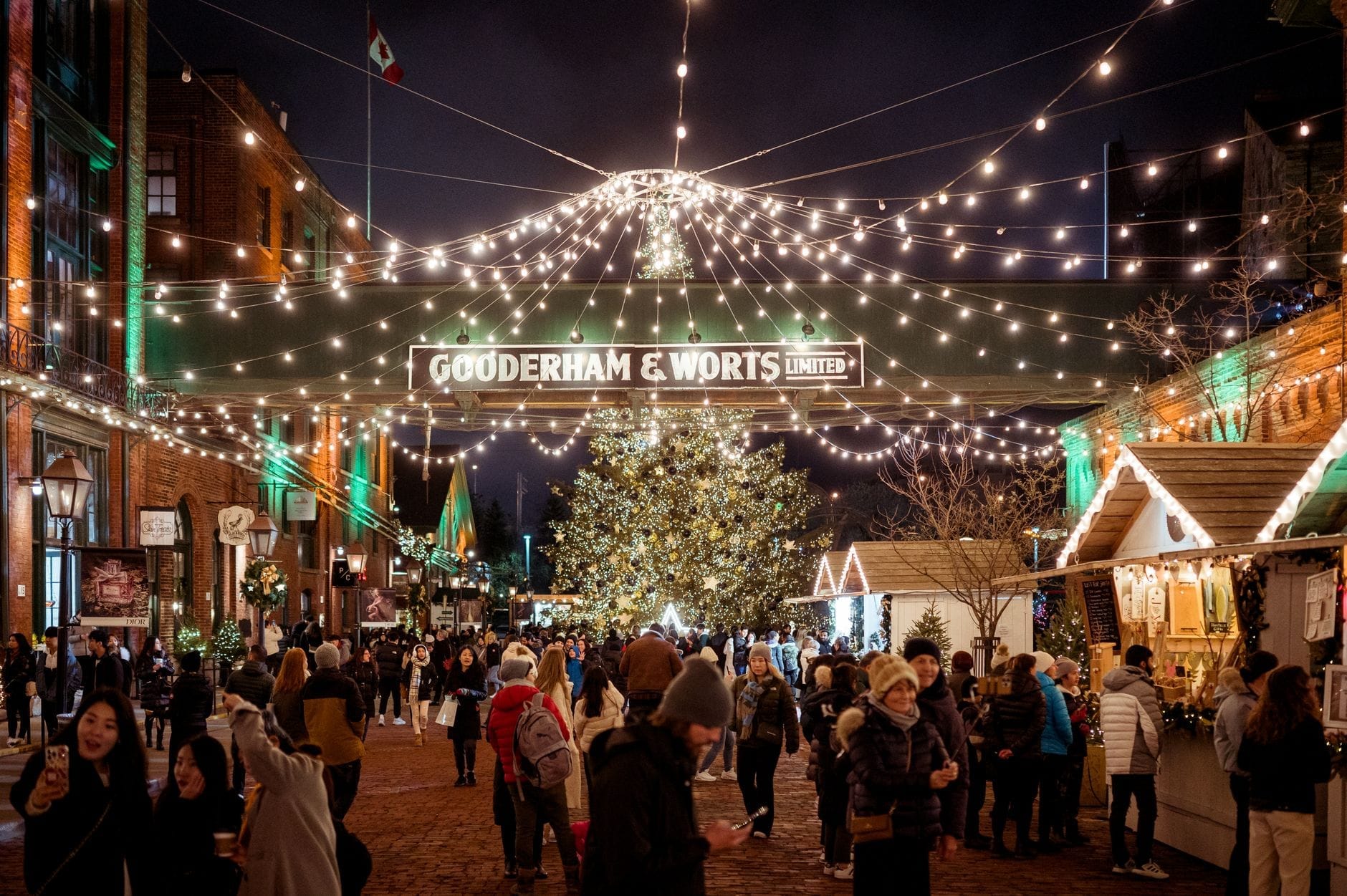 Cena festiva do mercado natalino no Distillery District de Toronto, com uma grande árvore de Natal, luzes decorativas, barracas de madeira e multidões aproveitando a noite.
