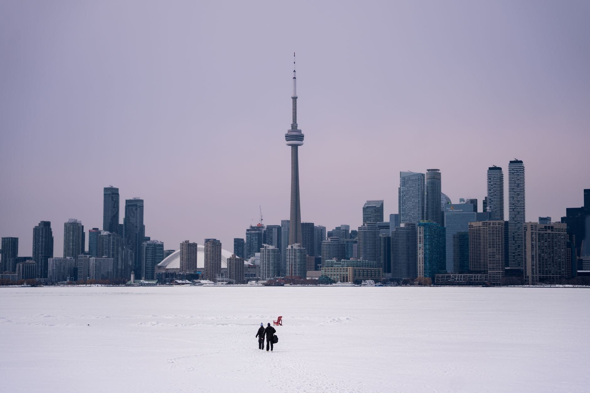 Duas pessoas caminhando sobre um lago congelado coberto de neve em direção ao horizonte de Toronto e à CN Tower sob um céu de inverno.