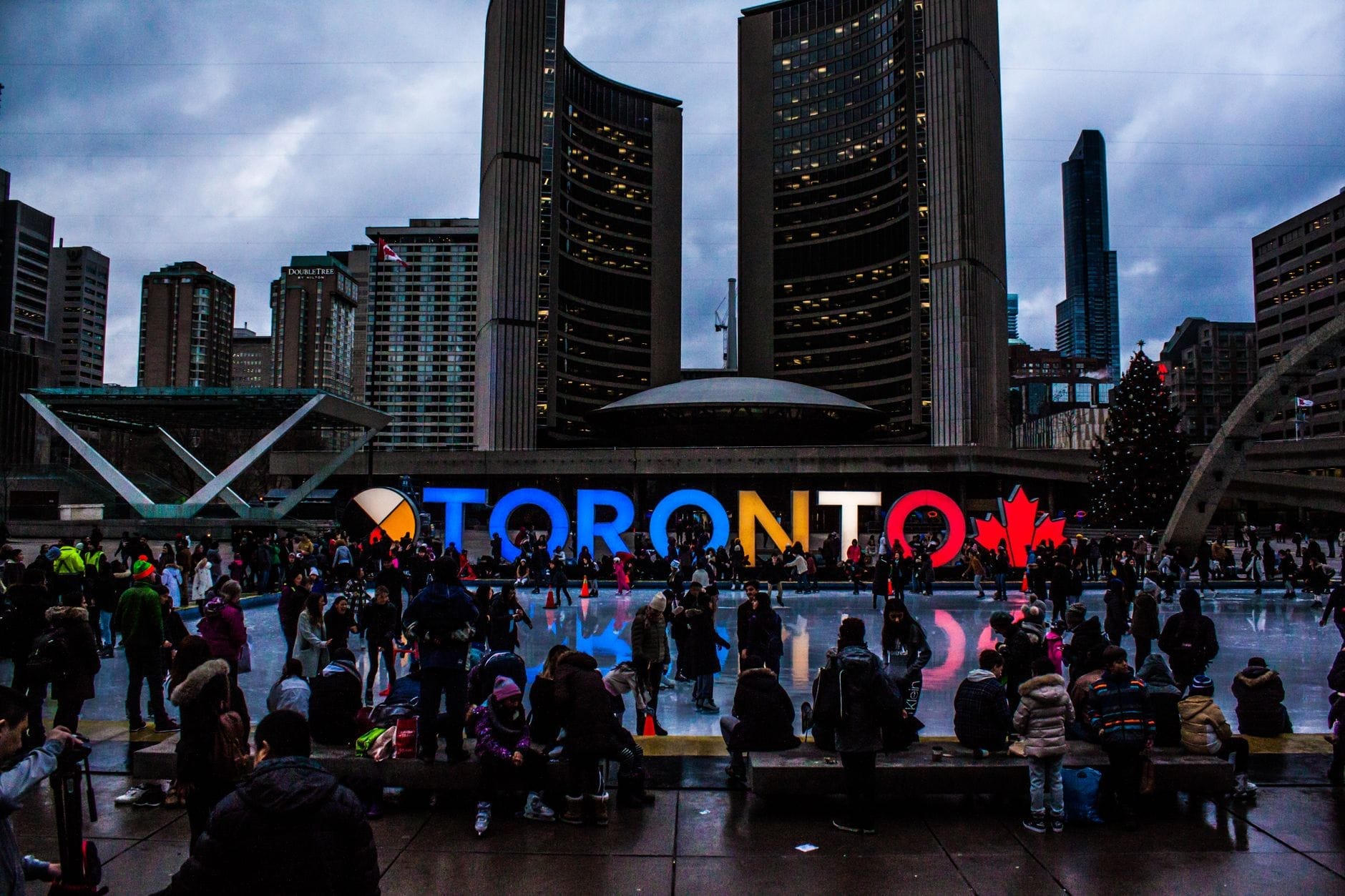 Multidão reunida em Nathan Phillips Square em frente à placa iluminada de TORONTO com os edifícios da prefeitura visíveis ao fundo.