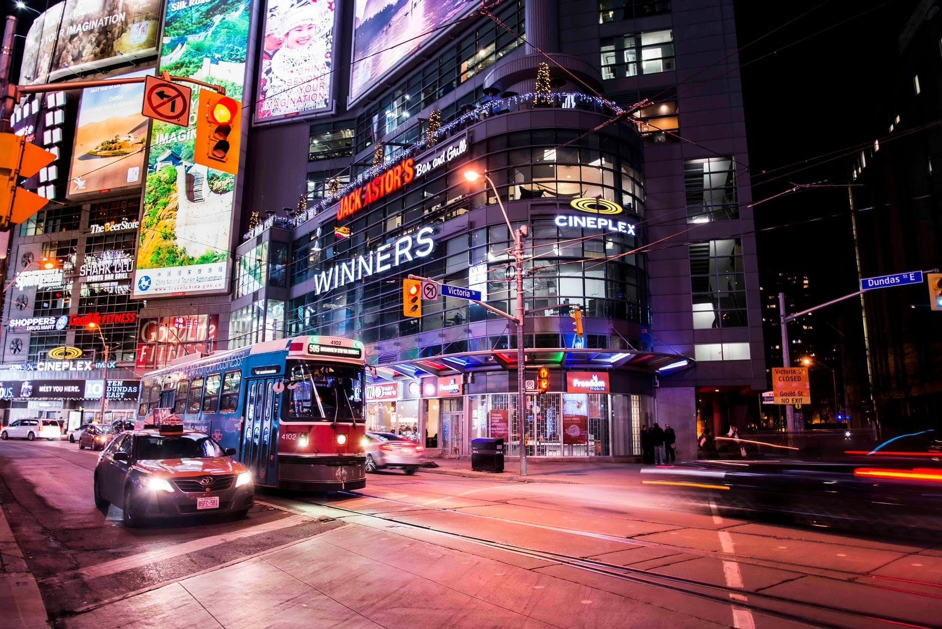Bonde passando pelo Cineplex e Winners em uma movimentada intersecção no centro de Toronto à noite, com outdoors iluminados e o agito da cidade.