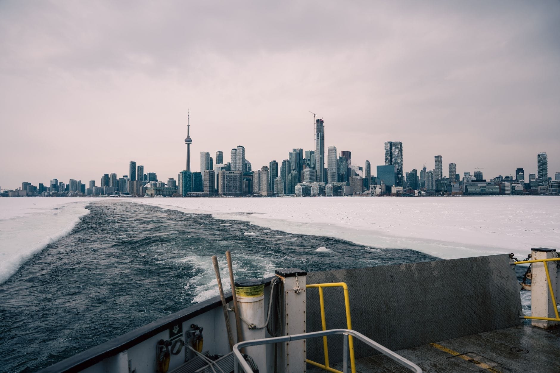 Vista da balsa com o skyline de Toronto no inverno, mostrando gelo no lago e céu nublado, destacando as condições fora de temporada.