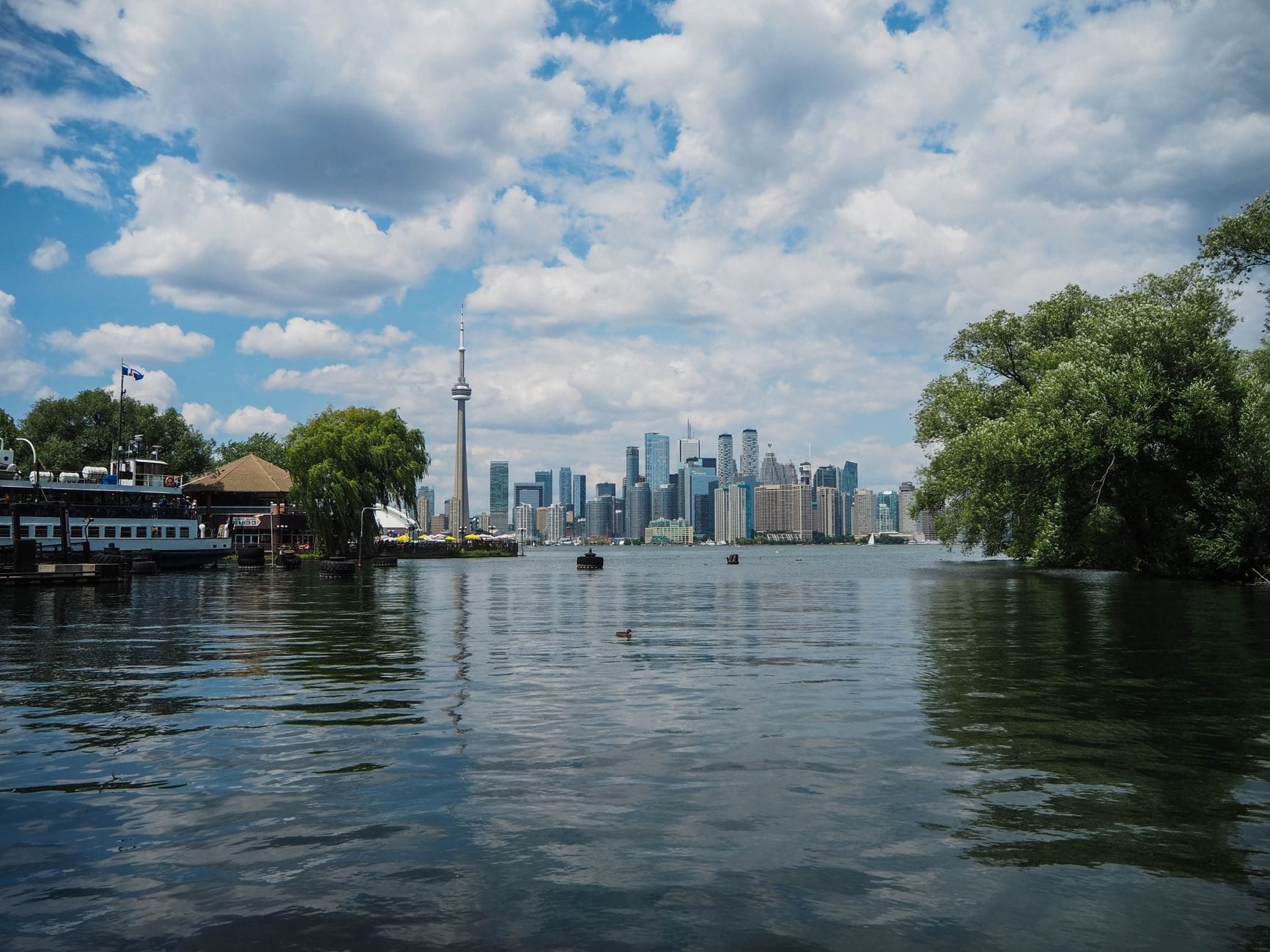 Vista das Ilhas Toronto mostrando árvores, água, um cais de balsa e o horizonte do centro de Toronto com a CN Tower em um dia parcialmente nublado.
