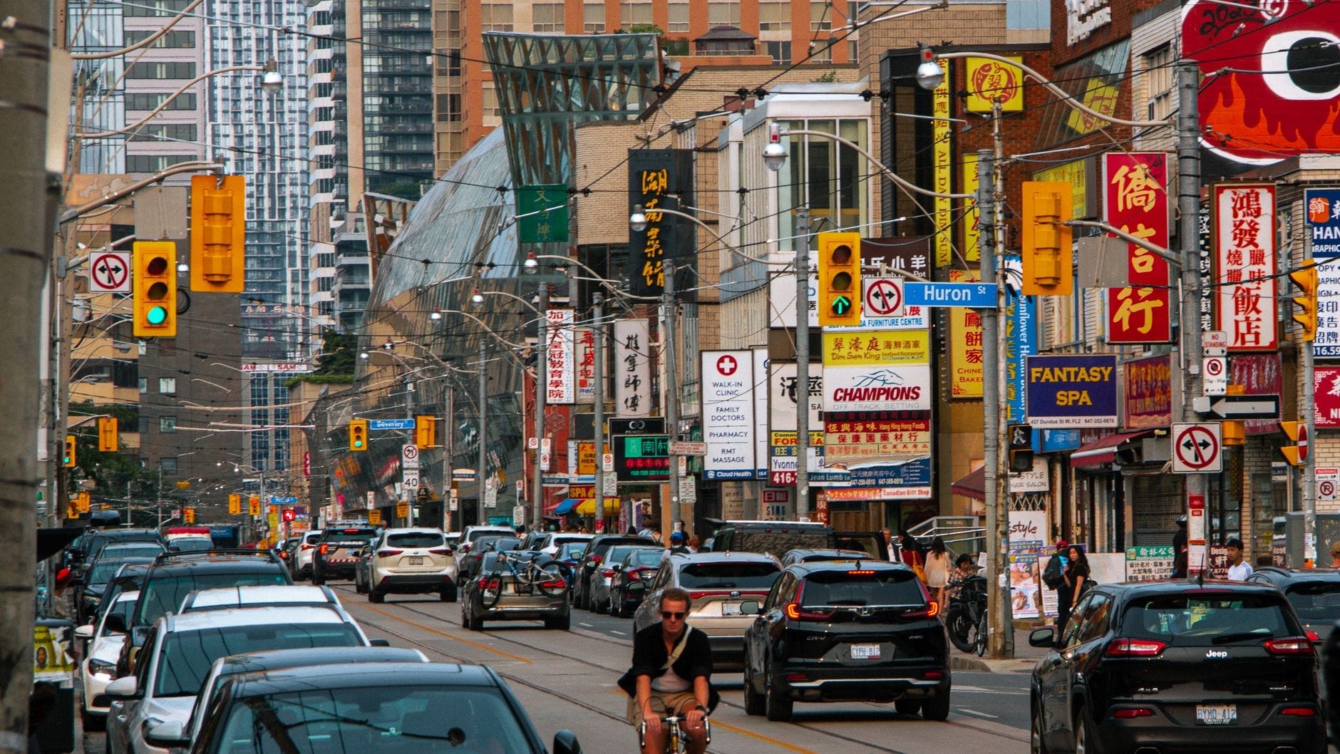 Rua movimentada de Toronto no distrito da Chinatown, cheia de carros, bicicletas e letreiros coloridos em vários idiomas, com os prédios modernos da cidade ao fundo.