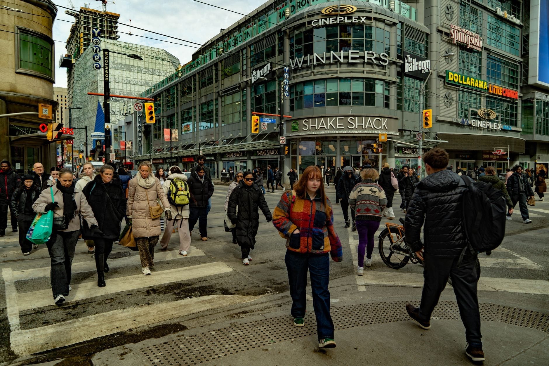 Movimentada interseção de Toronto com uma multidão diversificada atravessando em frente a lojas reconhecíveis e edifícios urbanos.
