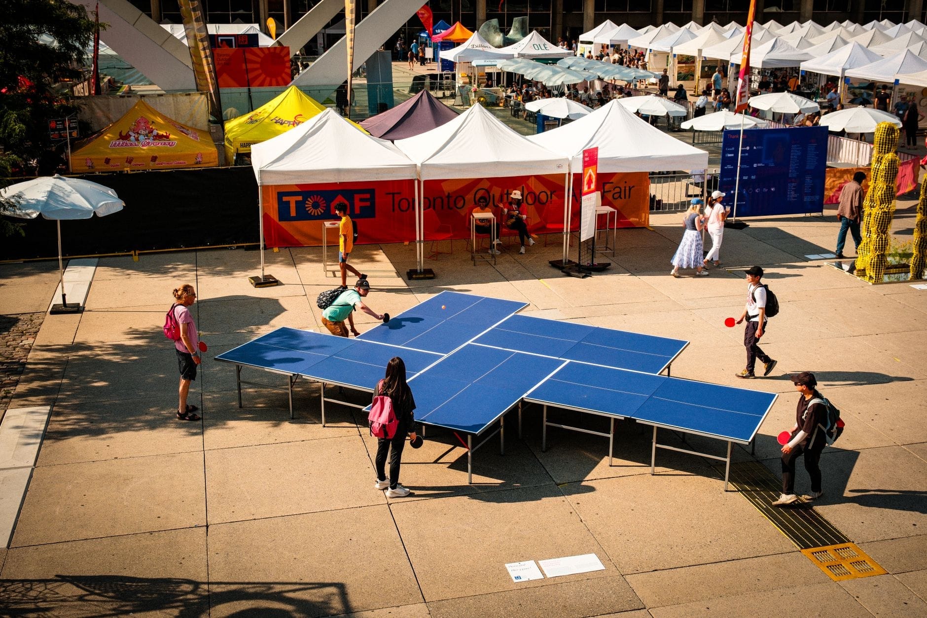 Pessoas jogando ping pong em uma feira de rua ao ar livre em Toronto, com tendas de festival e clima ensolarado de verão.