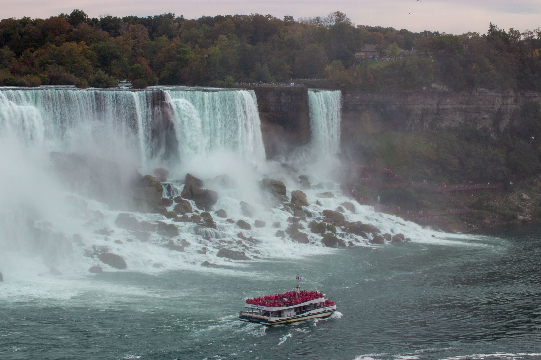 Vista ampla das Cataratas do Niágara com um barco turístico lotado de visitantes se aproximando da base das quedas e uma densa névoa se erguendo, cercado de árvores verdes.
