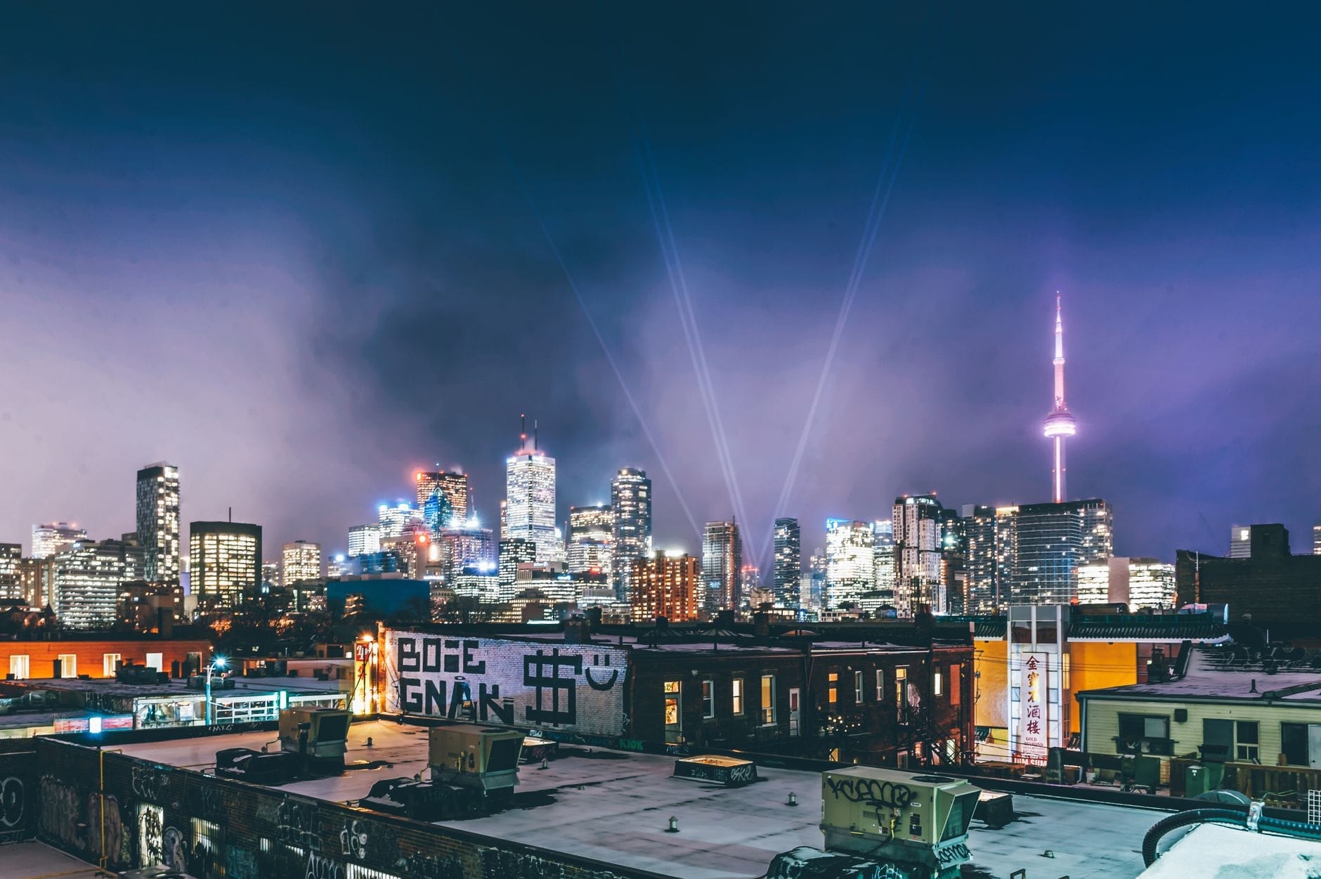 Horizonte de Toronto à noite com luzes brilhantes da cidade, a CN Tower iluminada e feixes de luz no céu sobre os telhados.