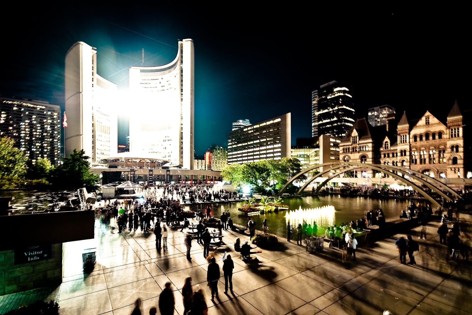 Multidões se reúnem na Nathan Phillips Square de Toronto à noite, com o City Hall brilhantemente iluminado e o espelho d'água rodeado de edifícios modernos e históricos.