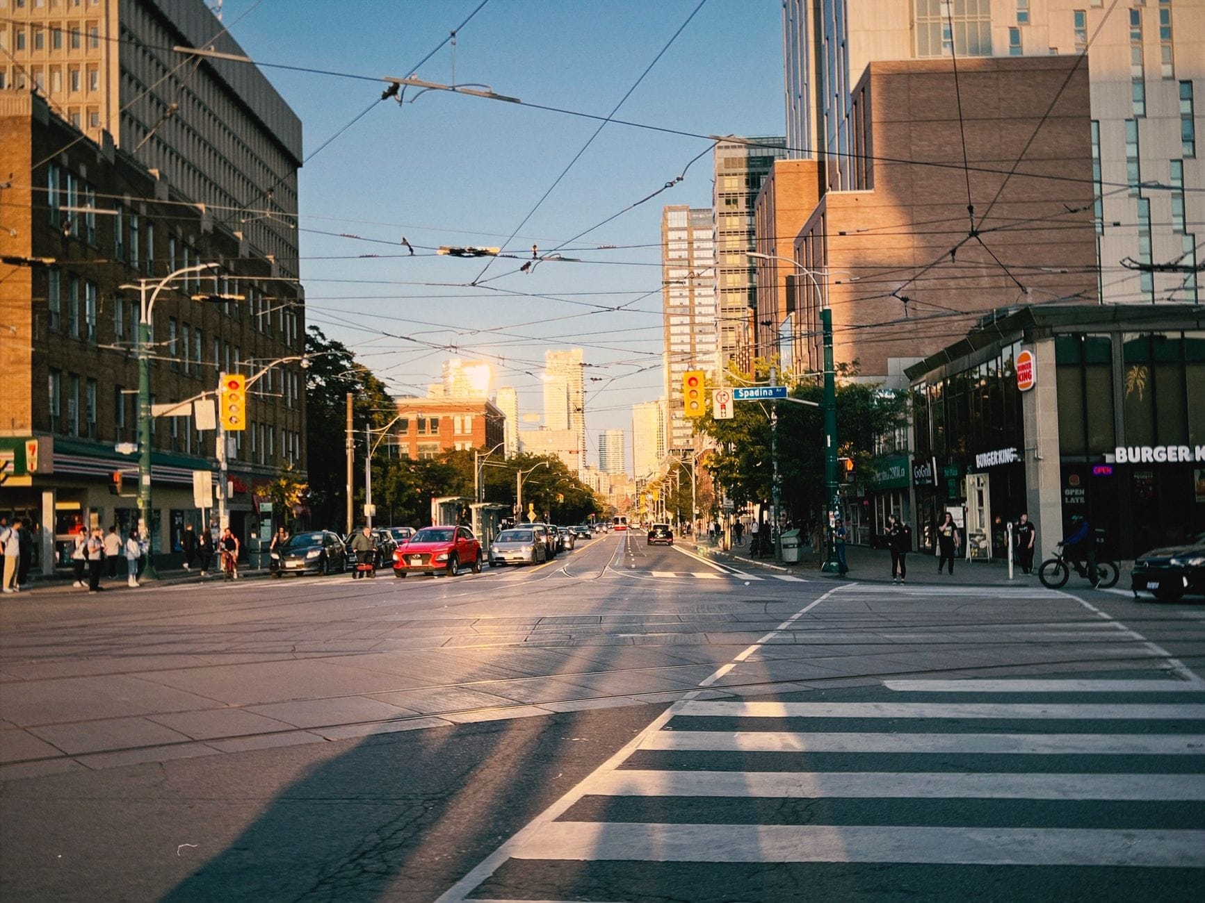 Vista ampla de uma movimentada rua de Toronto com restaurantes e locais de comida para viagem ao longo da calçada, fios elétricos acima e pessoas caminhando no final da tarde.
