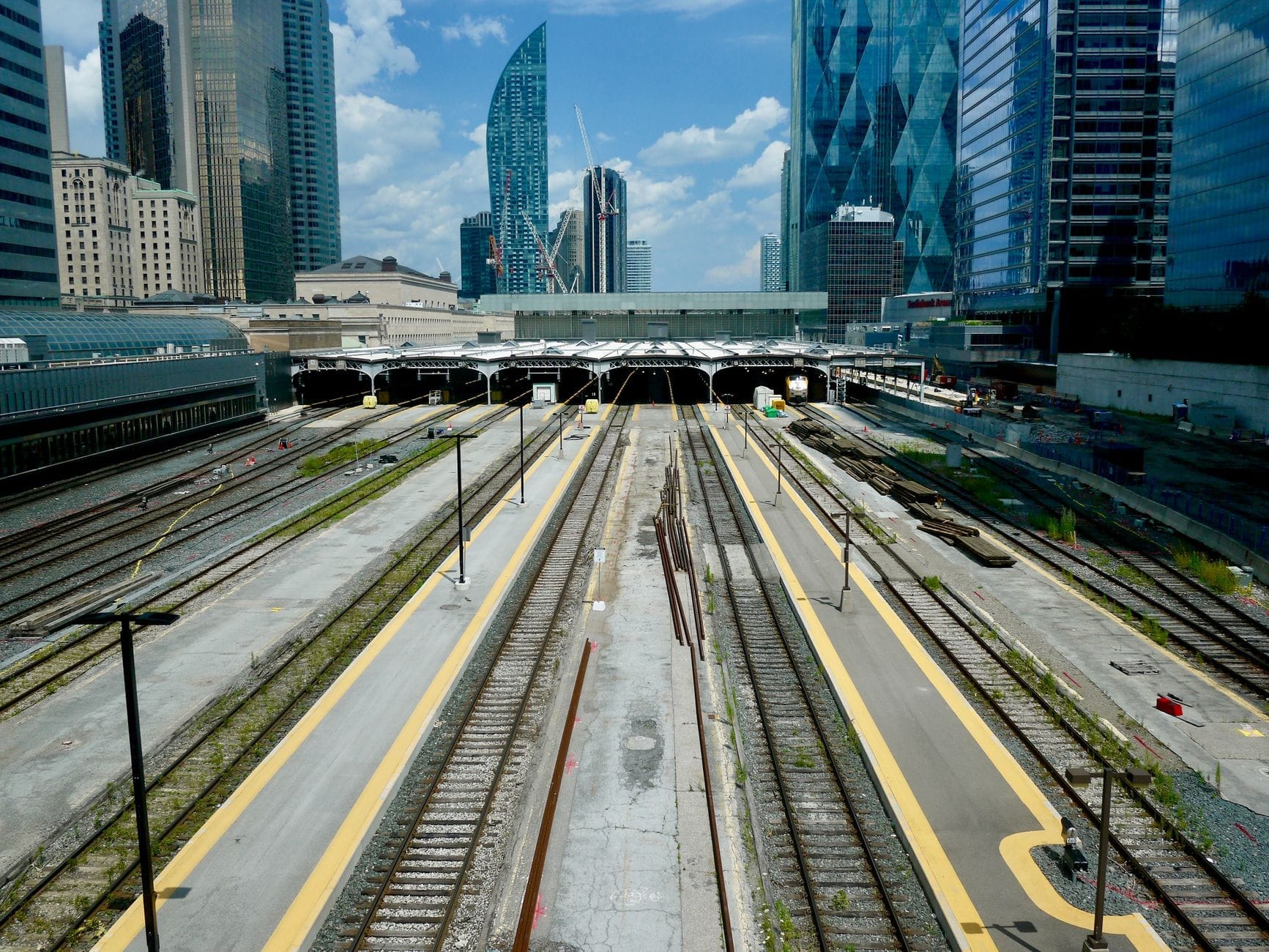 Vista dos trilhos ferroviários que levam à Toronto Union Station com os arranha-céus do centro ao fundo num dia ensolarado.