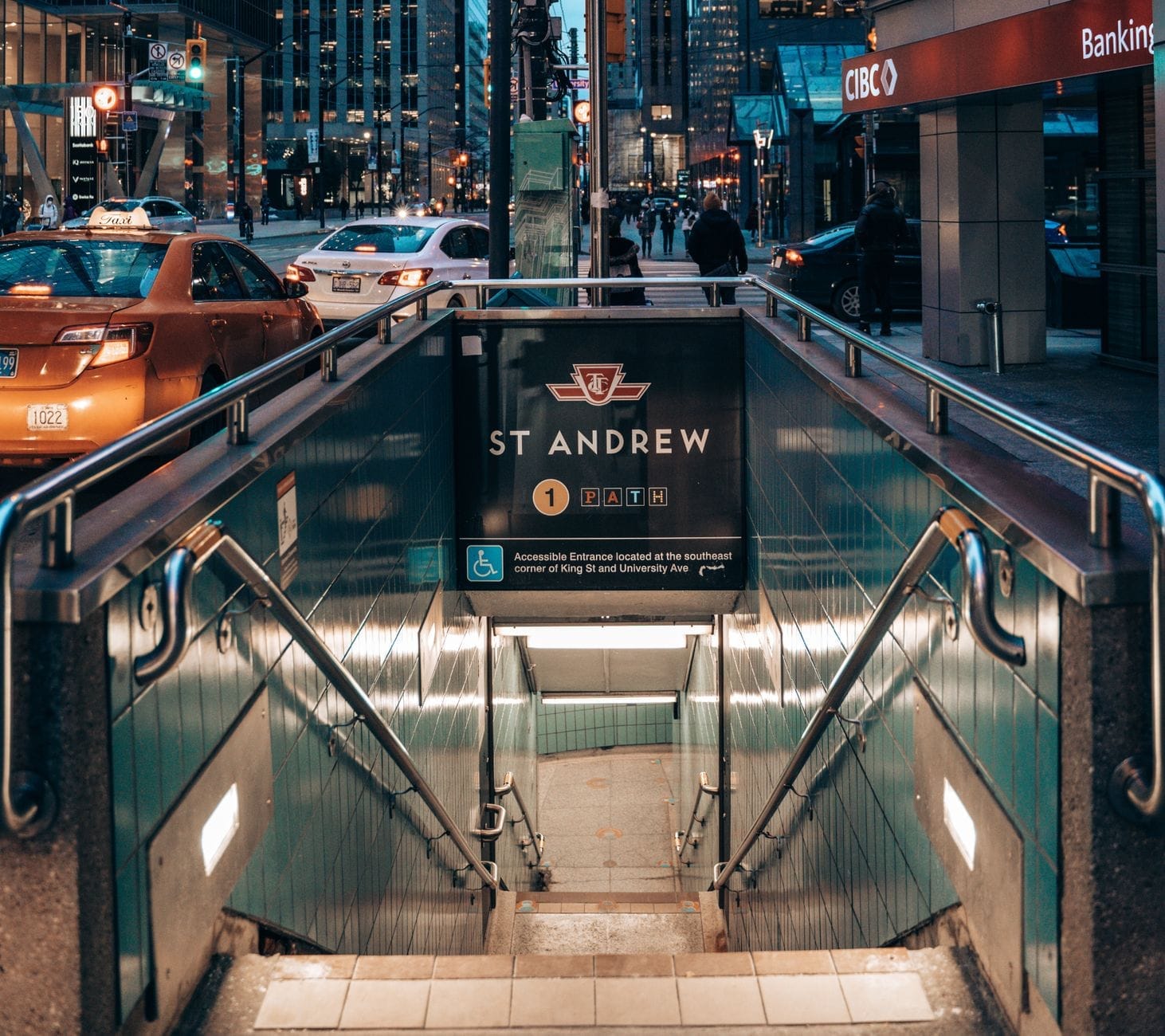 Entrada da estação de metrô St Andrew de Toronto ao nível da rua, com escadas descendo, cercada por edifícios da cidade e sinalização da TTC visível.