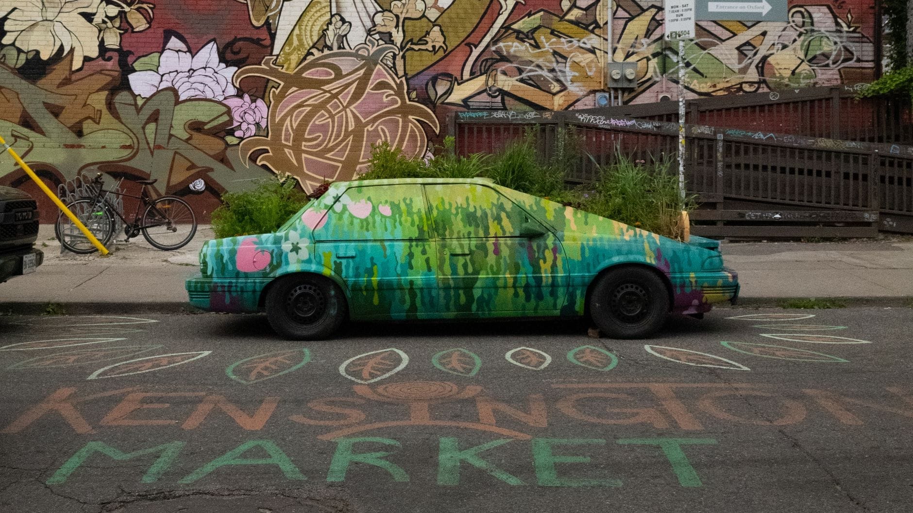Um carro pintado com cores vibrantes em frente a um mural numa rua animada com 'Kensington Market' pintado no asfalto em Toronto.