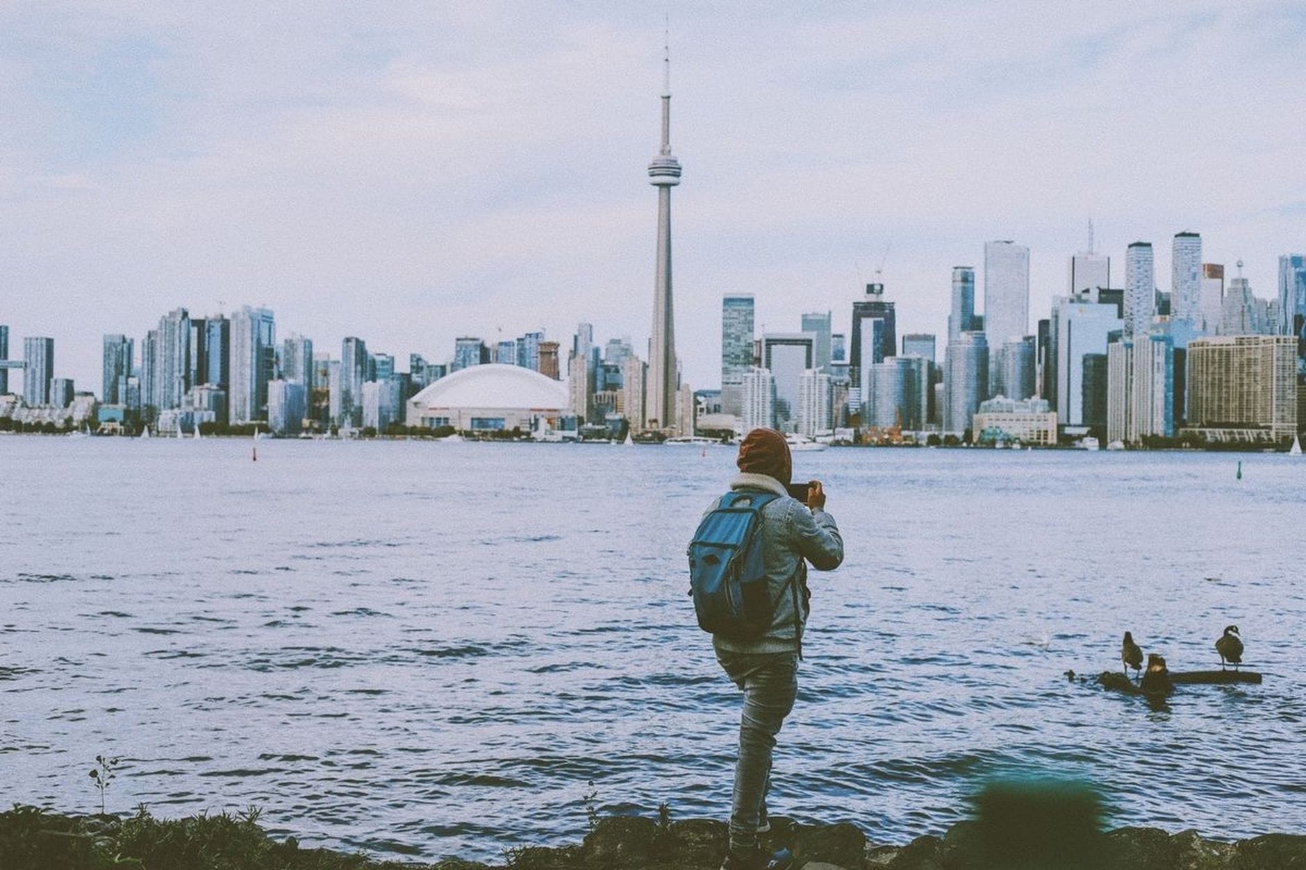 Viajante solo com mochila fotografando o skyline de Toronto do outro lado da água, com a CN Tower e os prédios do centro em um dia nublado.