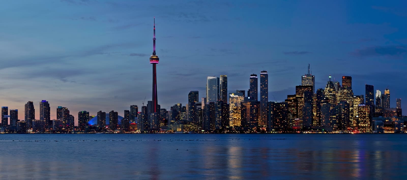 Toronto skyline at dusk with the CN Tower and city lights reflecting on Lake Ontario under a blue evening sky.