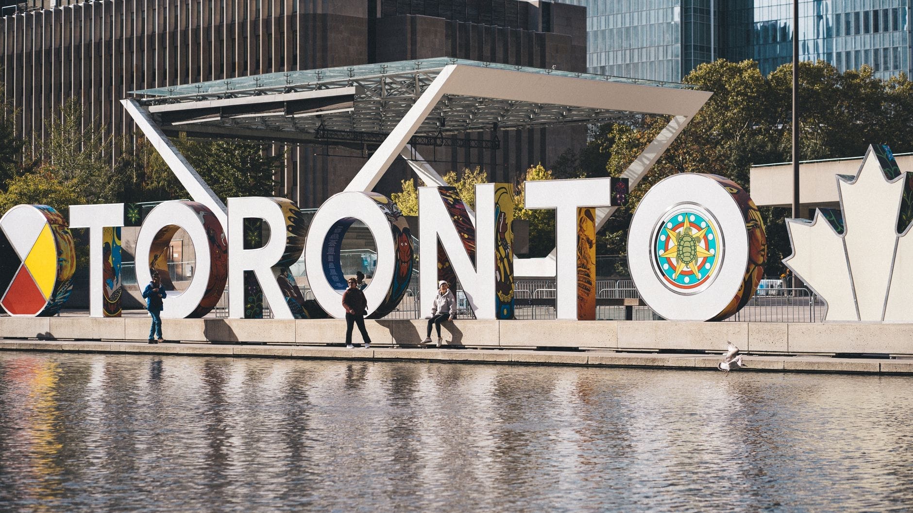 Des personnes marchant devant le célèbre panneau Toronto à Nathan Phillips Square, avec des bâtiments modernes en arrière-plan.