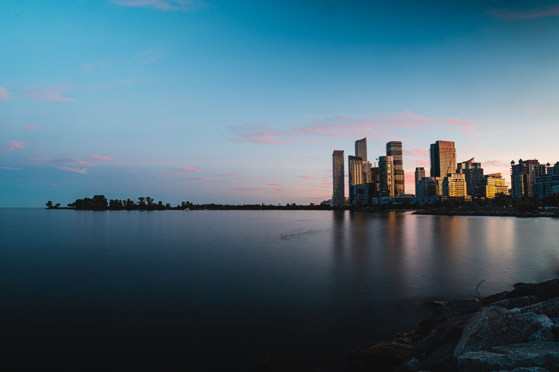Vista da orla de Toronto ao anoitecer com condomínios de alto padrão, áreas verdes e um lago calmo refletindo o céu, ilustrando o contraste da orla leste-oeste.
