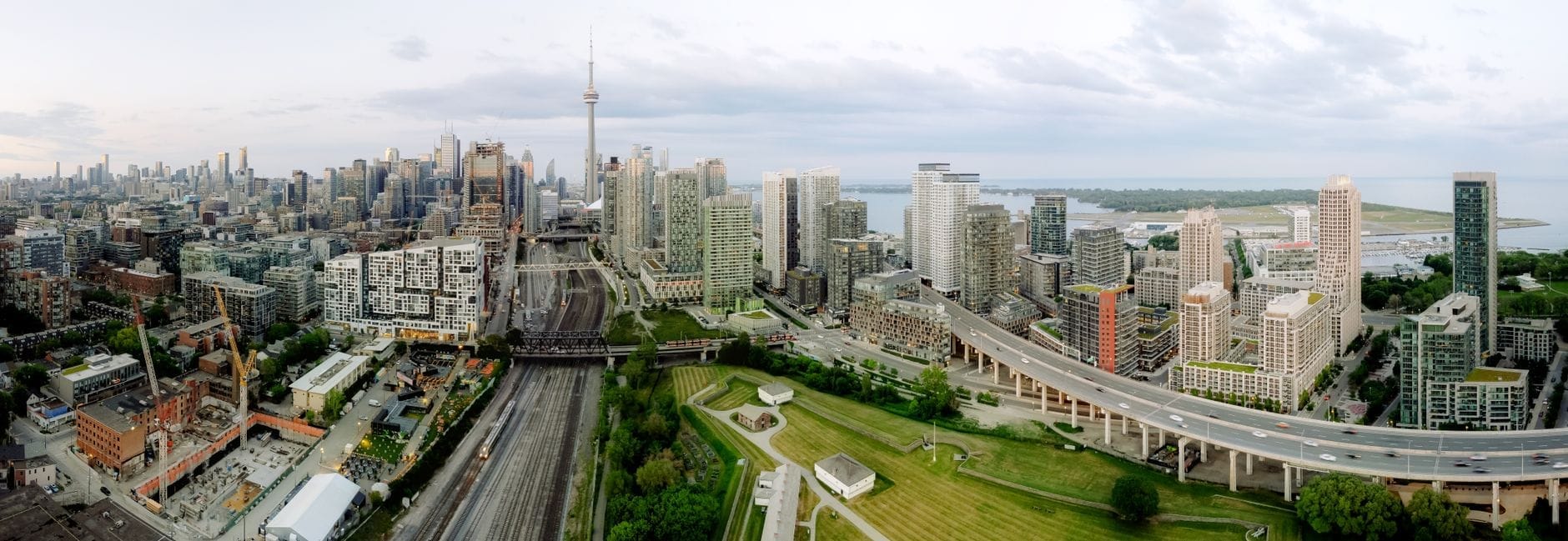 Vista aérea ampla do waterfront de Toronto, mostrando uma via expressa, espaços verdes, a orla do lago e o skyline com a CN Tower e edifícios altos.