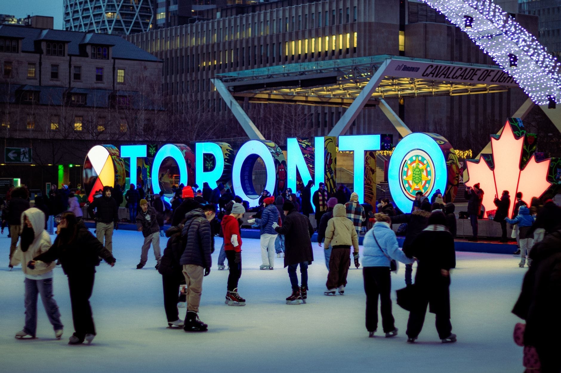 Pessoas de todas as idades patinando na Nathan Phillips Square em frente ao letreiro iluminado de Toronto, com os prédios da cidade ao fundo.