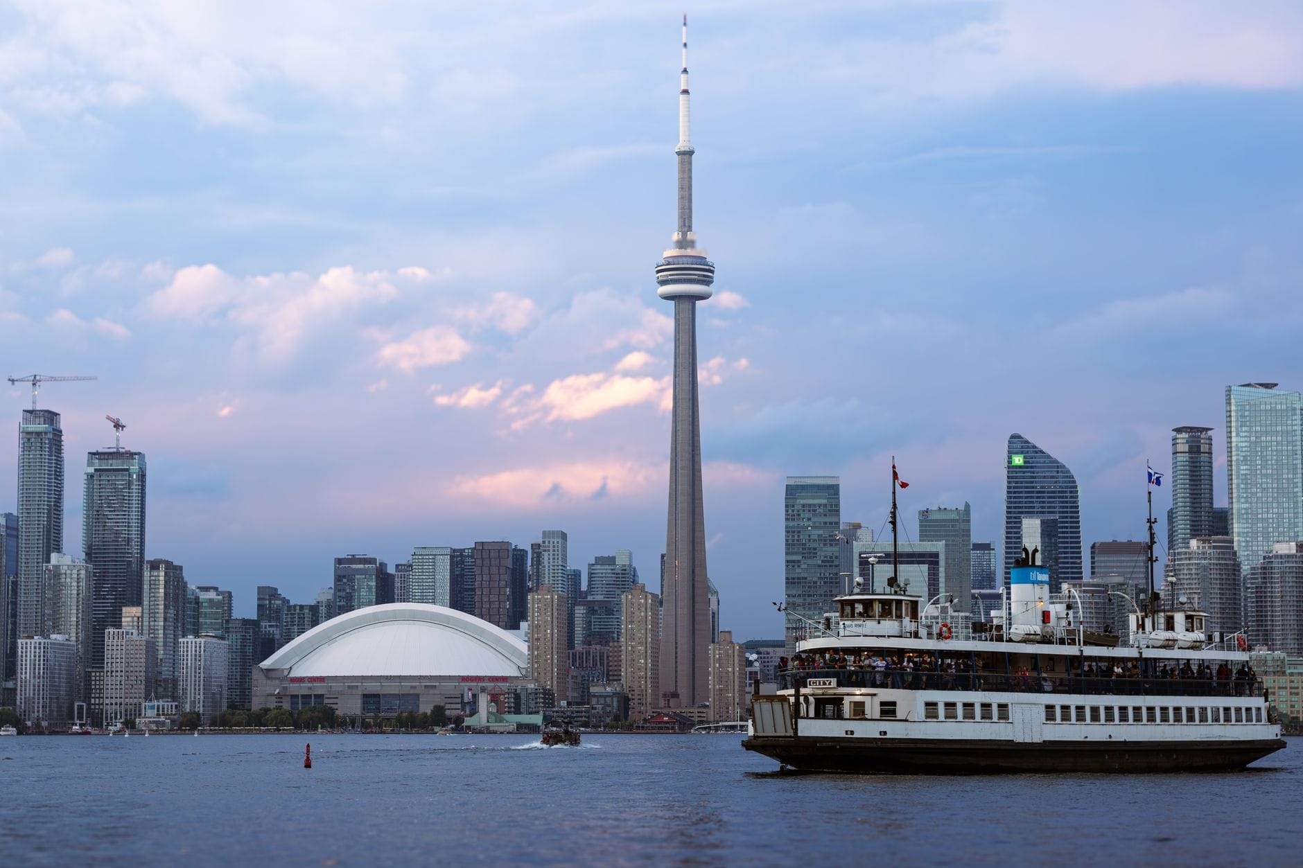 Horizonte de Toronto ao pôr do sol com a CN Tower, o Rogers Centre e uma balsa cruzando a orla marítima, destacando atrações icônicas para famílias.