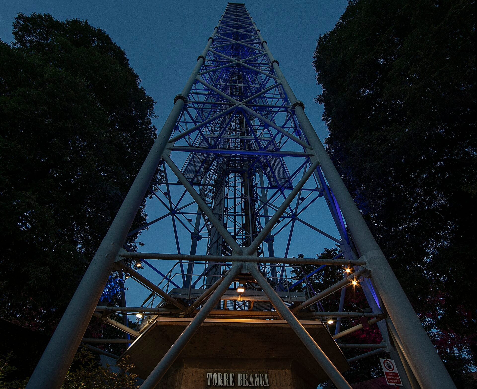 Vista dalla base della Torre Branca a Milano al crepuscolo, con struttura a traliccio in acciaio illuminata da luci blu e incorniciata da alberi alti.