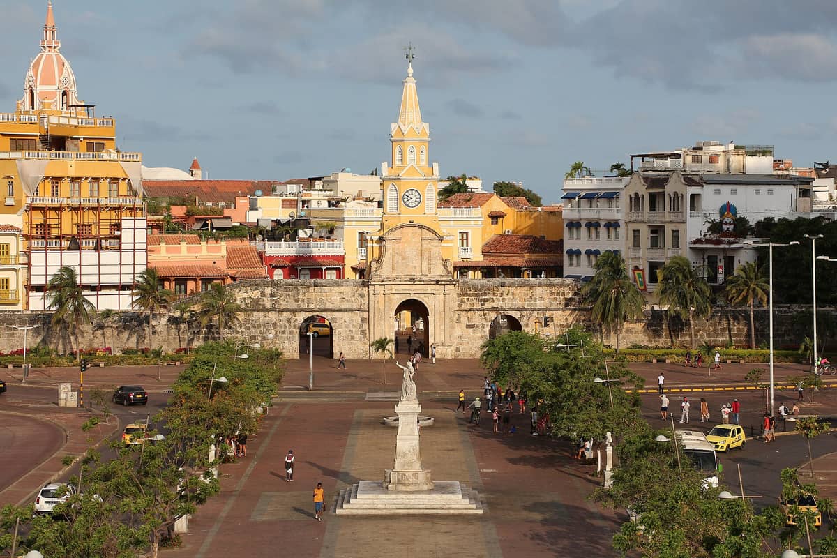 Front view of Cartagena's yellow Torre del Reloj (Clock Tower) at the gateway to the walled city, with arched entrance and plaza bustling with people.