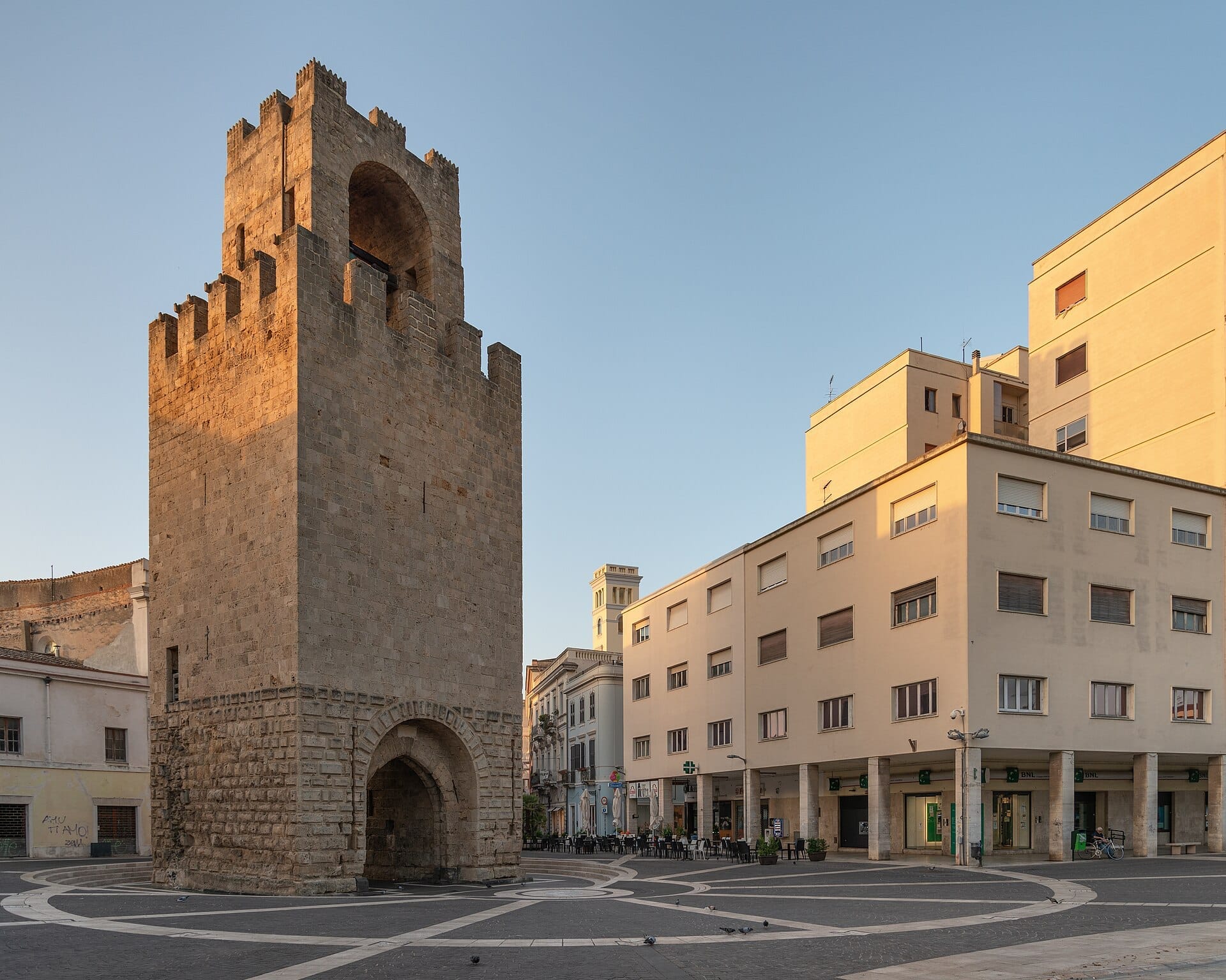 Der Torre di Mariano II ragt majestätisch auf der Piazza Roma in Oristano empor, umgeben von modernen Gebäuden im klaren Licht des frühen Abends.