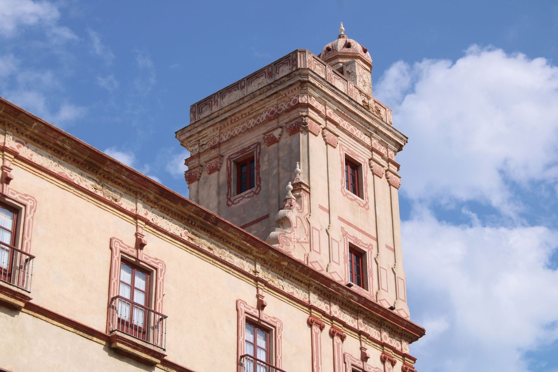 Torre Tavira watchtower rises above Cádiz, its ornate facade and rectangular tower contrasting with a bright blue sky and scattered clouds.