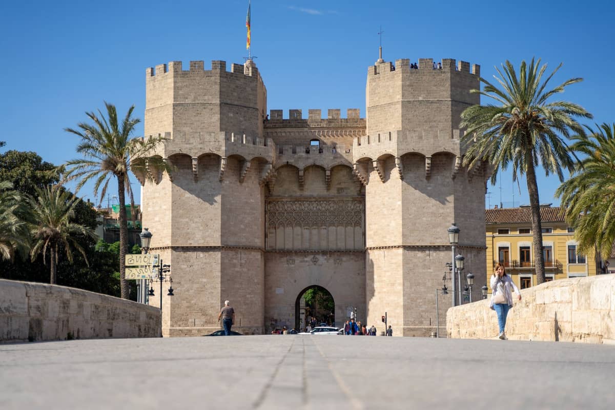 Vista ampia e nitida delle Torres de Serranos, le due torri medievali gemelle di Valencia, con palme e qualche passante sul ponte di pietra sotto un cielo azzurro.