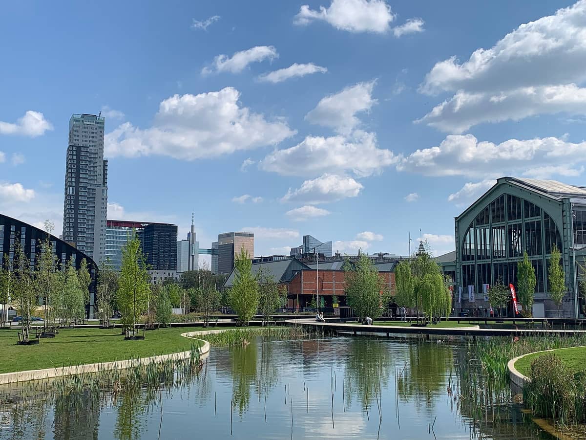 Tour & Taxis Bruxelas com históricos edifícios industriais, modernos arranha-céus ao fundo, parque ajardinado e lago refletivo sob um céu azul brilhante.