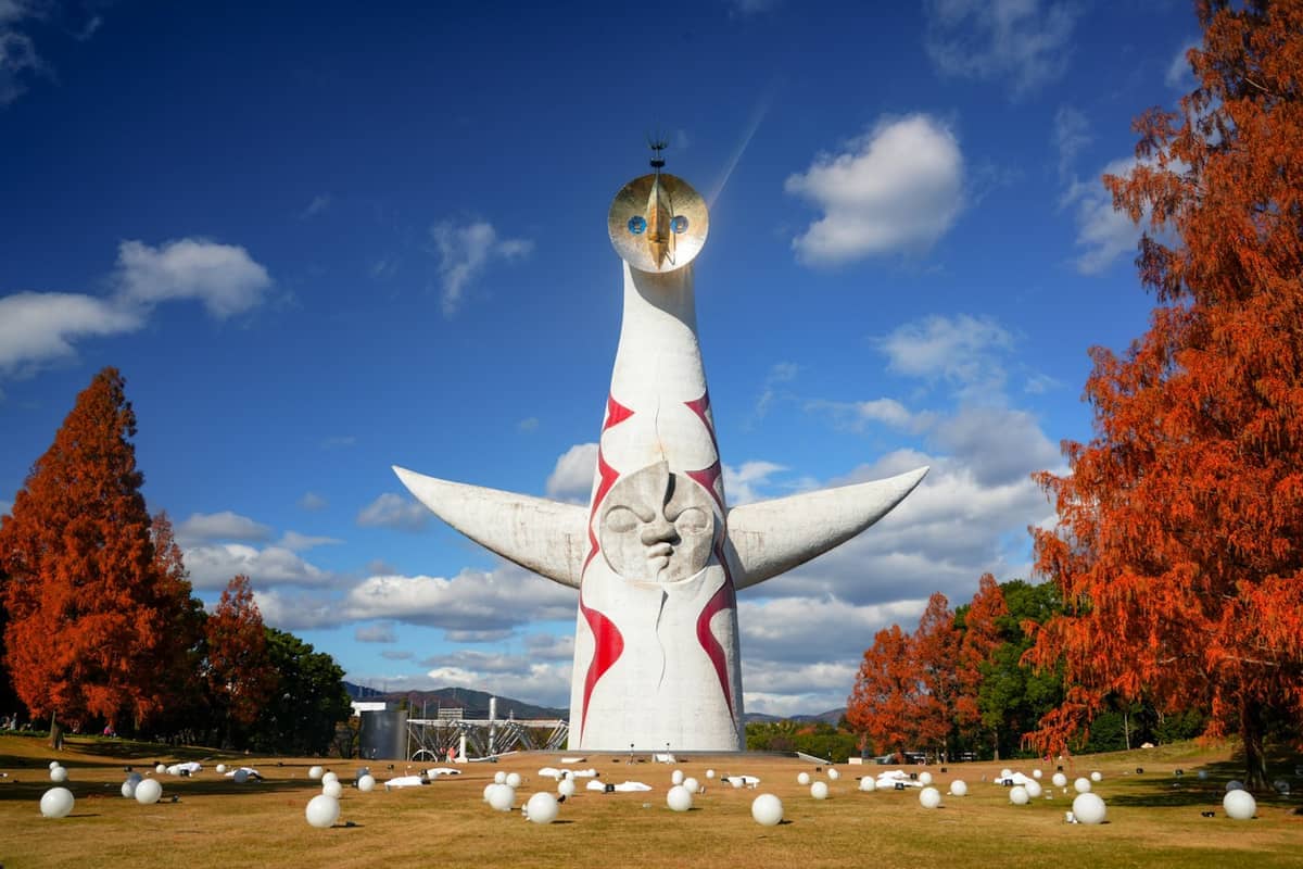 The Tower of the Sun sculpture rises on a grassy lawn surrounded by orange autumn trees and scattered white orbs under a blue sky in Osaka's Expo Park.