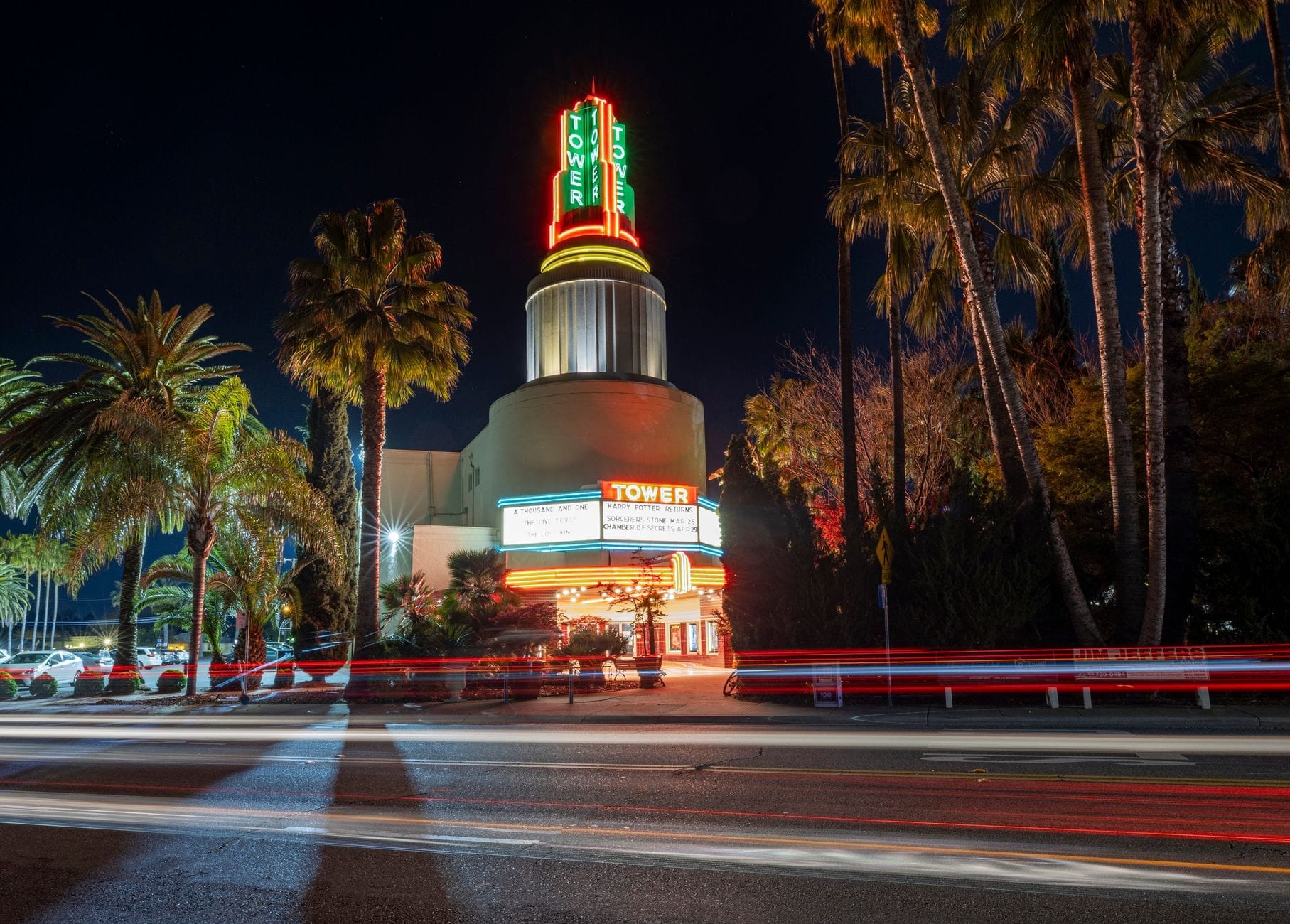 Vista notturna di un cinema art déco con una torre al neon illuminata con la scritta TOWER, circondata da palme e scie di luci su una strada animata.