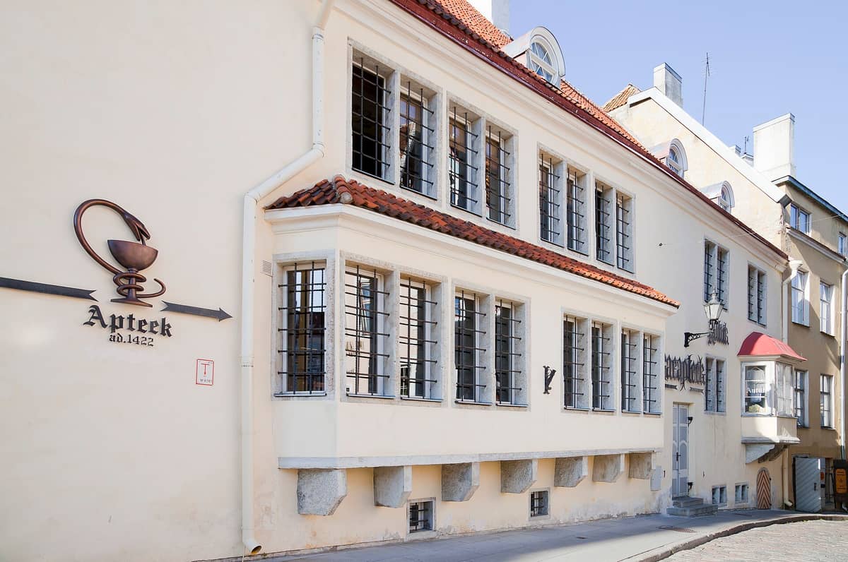 Die Außenfassade der Ratsapteek (Raeapteek) in Tallinn, Estland: cremefarben verputzt, mit vergitterten Fenstern und einem Schild mit der Aufschrift „Apteek ad. 1422".