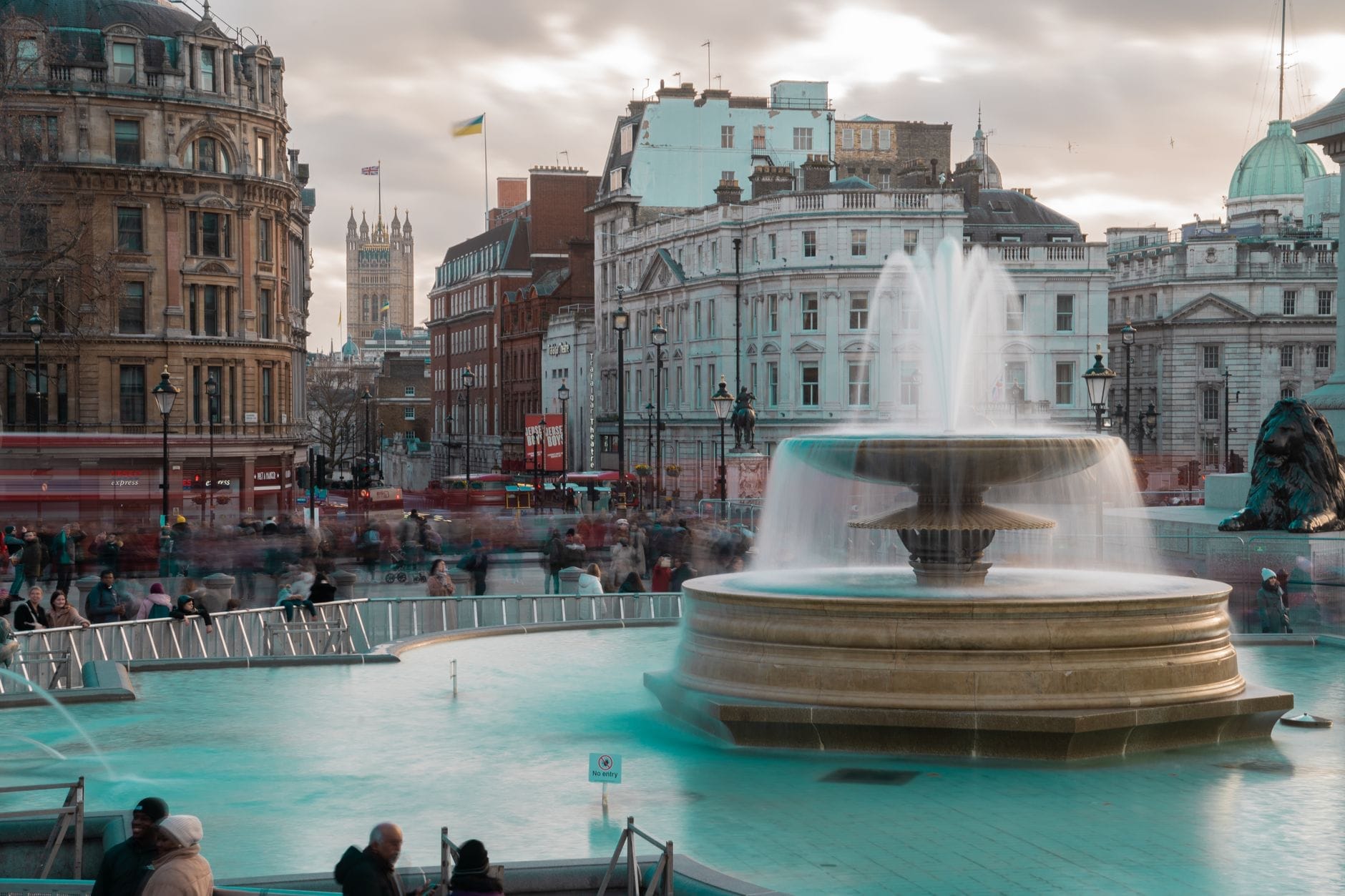 Vista di Trafalgar Square con la fontana centrale, gli edifici storici circostanti e la folla di visitatori in una giornata nuvolosa a Londra.