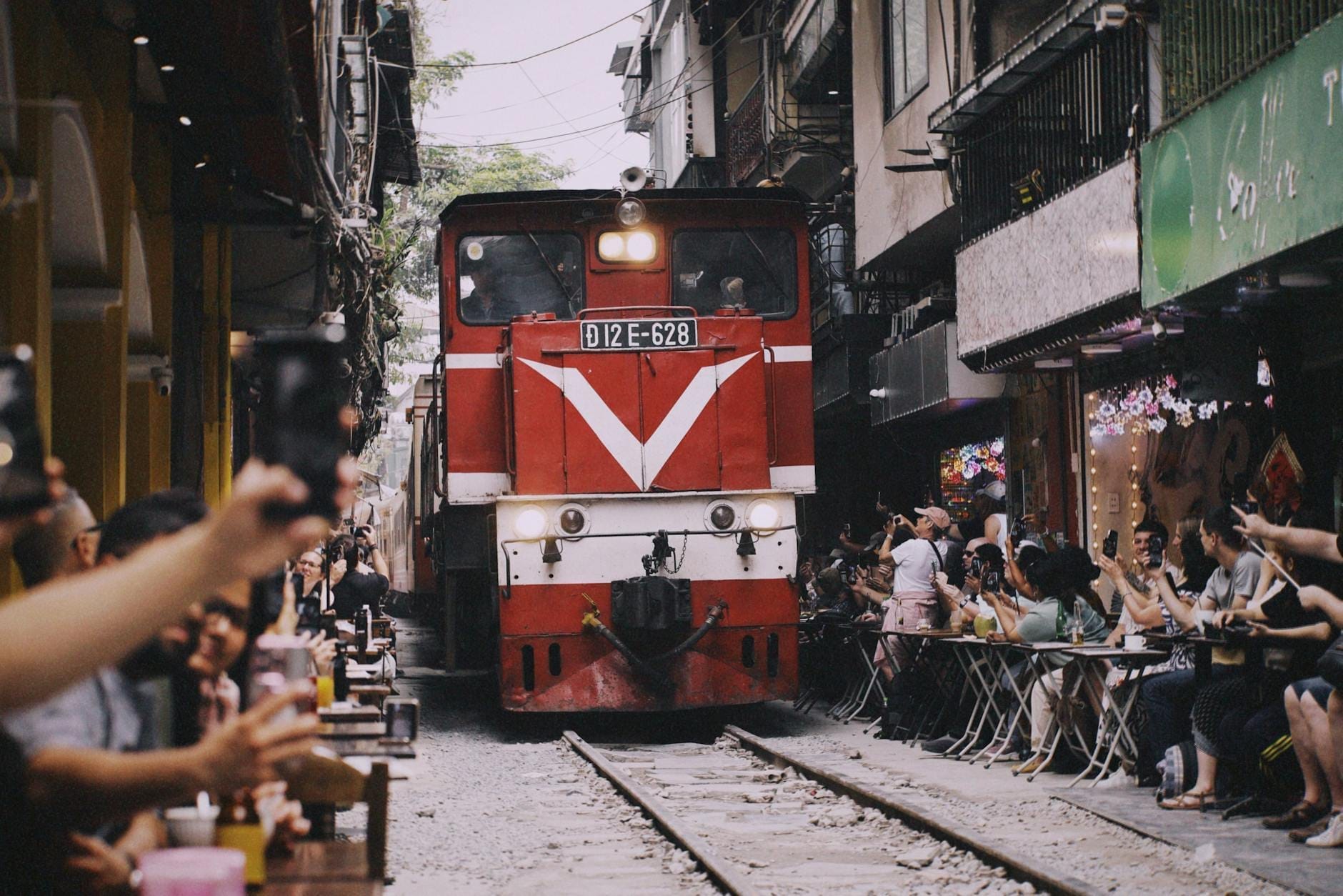 A red train passes through a narrow residential alley in Hanoi, closely flanked by buildings and outdoor cafes with people watching and taking photos.