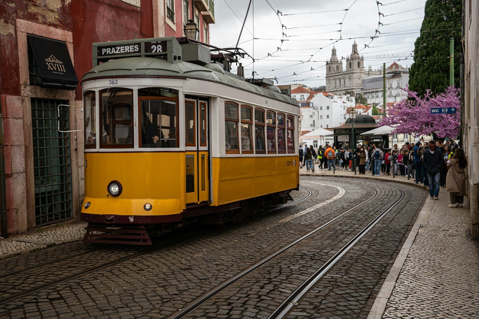 Il classico tram giallo 28E percorre una curva su un vicolo di sampietrini a Lisbona, con vecchi edifici, fiori primaverili e un gruppo di turisti sullo sfondo.