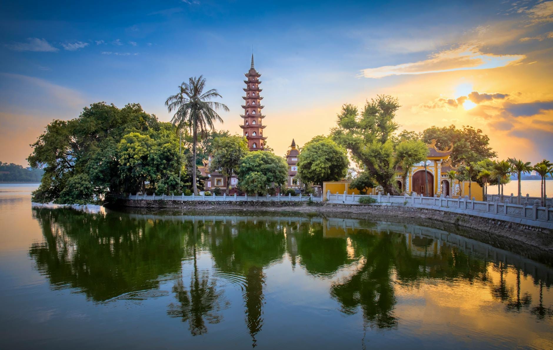 Tran Quoc Pagoda on a small island in West Lake at sunset, with lush trees and still water reflecting the historic pagoda, vibrant sky, and entrance gate.