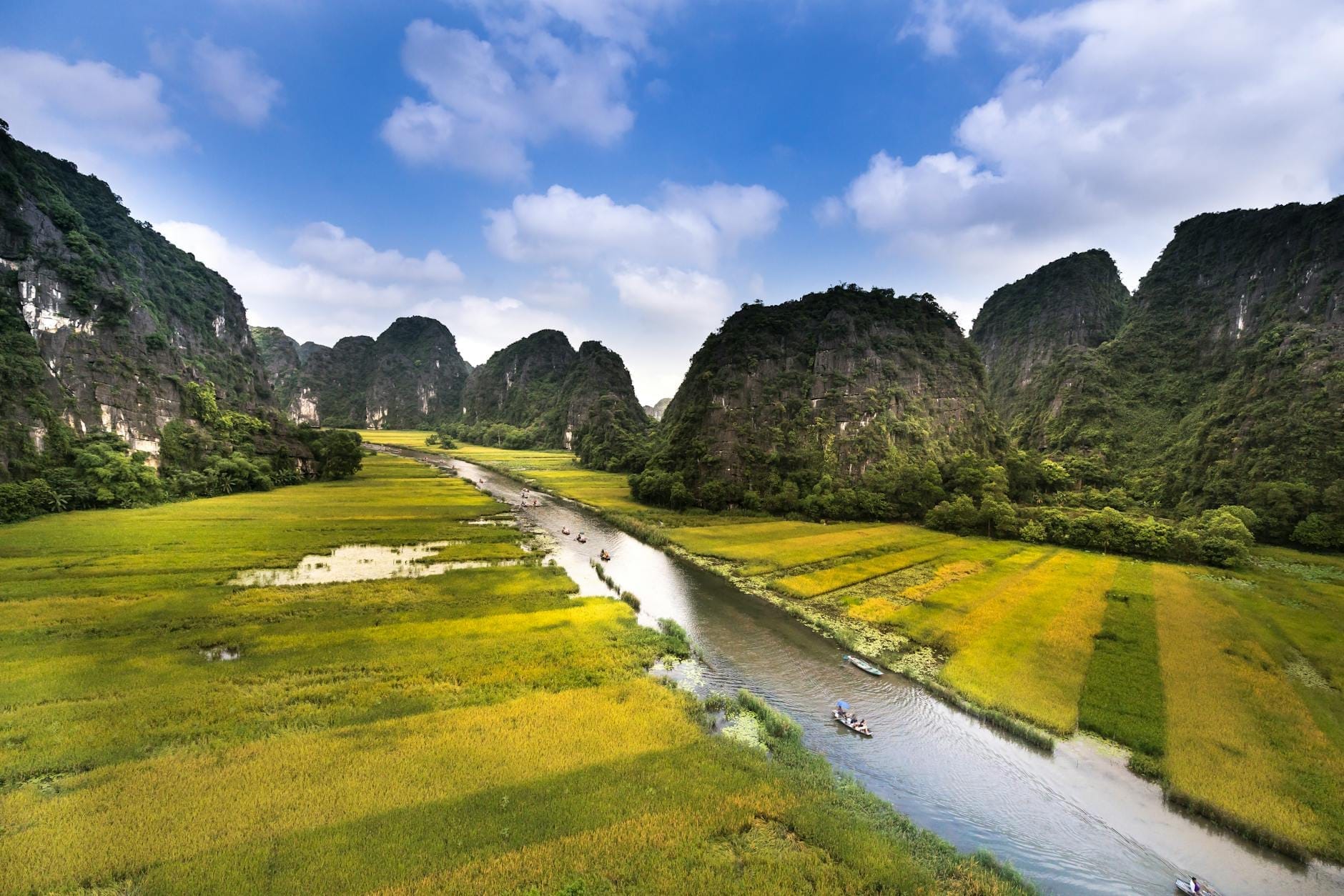 A scenic aerial view of the Trang An river winding through lush green fields and dramatic limestone karst mountains under a blue sky with scattered clouds.